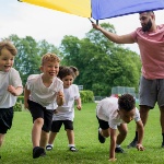 Four children in white t-shirts and black shorts are running and playing underneath a multi-coloured tarp held by an adult. 